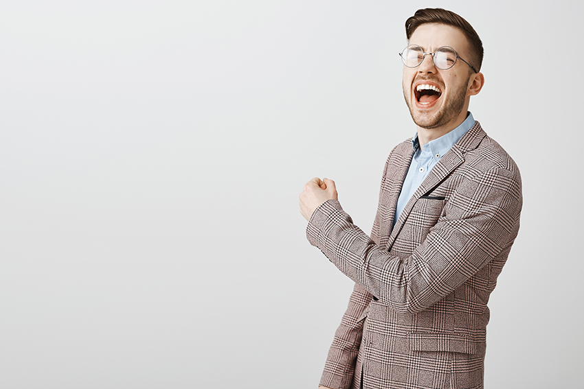 Handsome pleased caucasian male in jacket and glasses yelling from triumph and joy raising fist in victory or success gesture over gray wall.